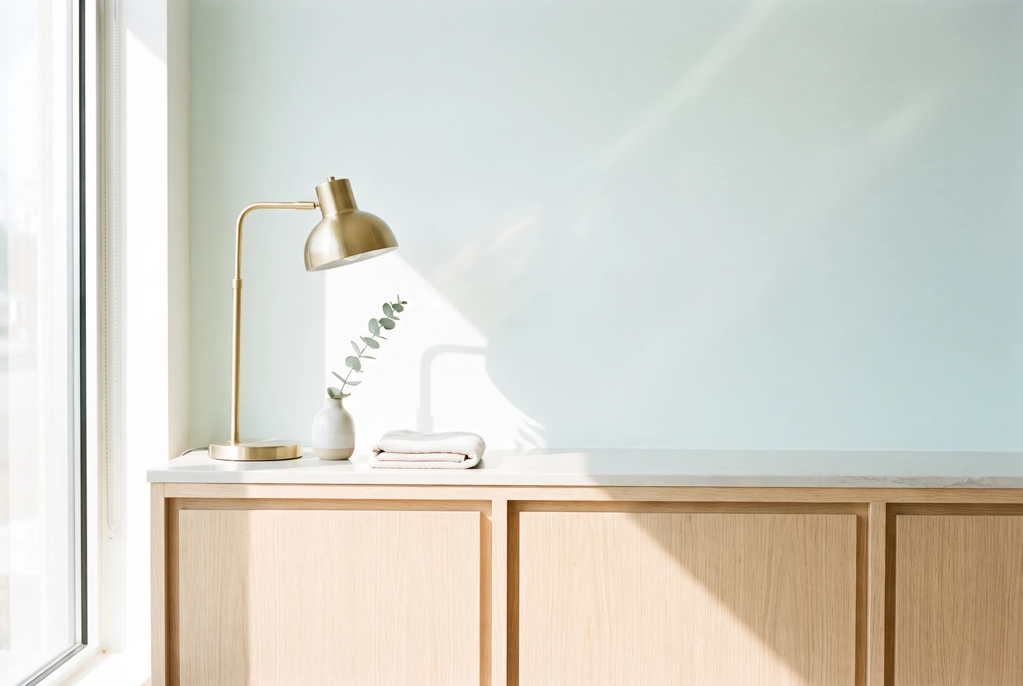 A bright reception counter in pale oak and stone with a brass lamp and a single eucalyptus stem